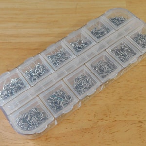 May include: A clear plastic storage box with 12 compartments filled with small silver jump rings.