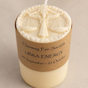 Könnte beinhalten: Eine cremefarbene zylindrische Kerze mit einem weißen Docht. Die Oberseite zeigt ein erhabenes Relief einer Waage. Ein braunes Papieretikett umschließt die Mitte mit dem Text: Charming. Fair. Sociable. LIBRA ENERGY. 23 September - 22 October.