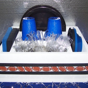 May include: White plastic serving tray with a black and white football themed ribbon. The tray has a black plastic plate, two blue plastic cups, and a bundle of clear plastic forks and spoons.