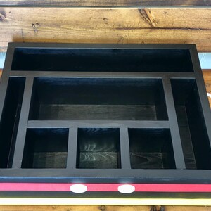 Puede incluir: Organizador de madera negro con m&uacute;ltiples compartimentos y asas blancas. El organizador tiene un dise&ntilde;o de rayas rojas y amarillas en la parte delantera con dos acentos circulares blancos. El interior est&aacute; dividido en varias secciones de almacenamiento.