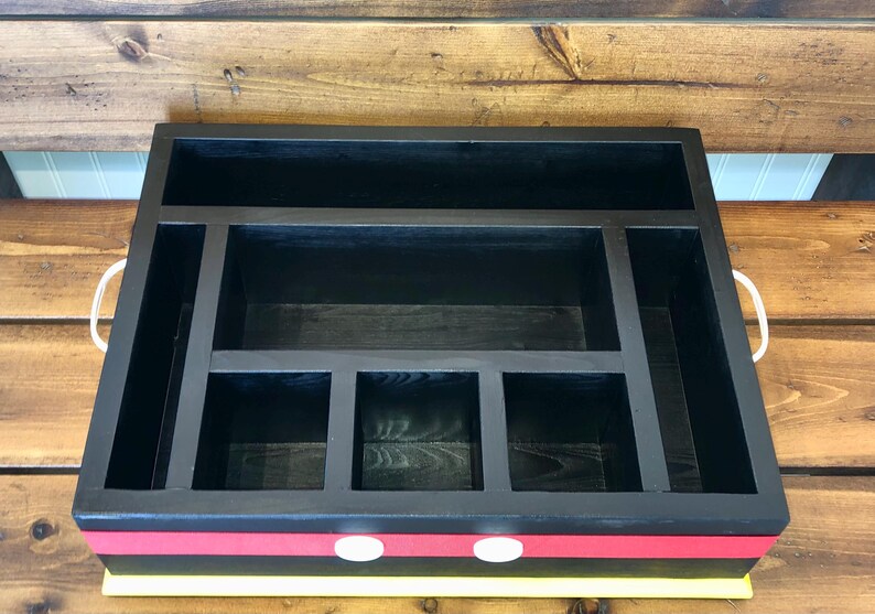 Puede incluir: Organizador de madera negro con m&uacute;ltiples compartimentos y asas blancas. El organizador tiene un dise&ntilde;o de rayas rojas y amarillas en la parte delantera con dos acentos circulares blancos. El interior est&aacute; dividido en varias secciones de almacenamiento.