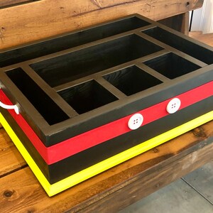 Puede incluir: Un organizador de madera negro con m&uacute;ltiples compartimentos. Cuenta con una franja roja, ribete amarillo y detalles de botones blancos. Un asa blanca est&aacute; en el lateral. El organizador est&aacute; sobre un banco de madera.