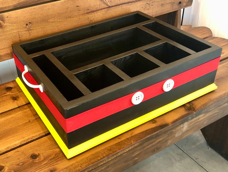 Puede incluir: Un organizador de madera negro con m&uacute;ltiples compartimentos. Cuenta con una franja roja, ribete amarillo y detalles de botones blancos. Un asa blanca est&aacute; en el lateral. El organizador est&aacute; sobre un banco de madera.