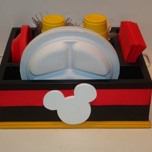 Puede incluir: Un organizador de madera negro y rojo con un dise&ntilde;o de cabeza de Mickey Mouse blanco. El organizador contiene platos blancos divididos, vasos amarillos, cuencos rojos, servilletas rojas y cubiertos de pl&aacute;stico transparente. La base es amarilla y tiene asas plateadas.