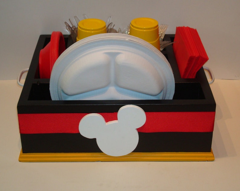Puede incluir: Un organizador de madera negro y rojo con un dise&ntilde;o de cabeza de Mickey Mouse blanco. El organizador contiene platos blancos divididos, vasos amarillos, cuencos rojos, servilletas rojas y cubiertos de pl&aacute;stico transparente. La base es amarilla y tiene asas plateadas.