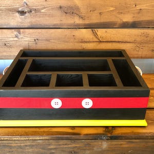 Puede incluir: Un organizador de madera negro con m&uacute;ltiples compartimentos. Presenta una franja roja con detalles de botones blancos y una base amarilla. El organizador est&aacute; dise&ntilde;ado para el almacenamiento y la organizaci&oacute;n.