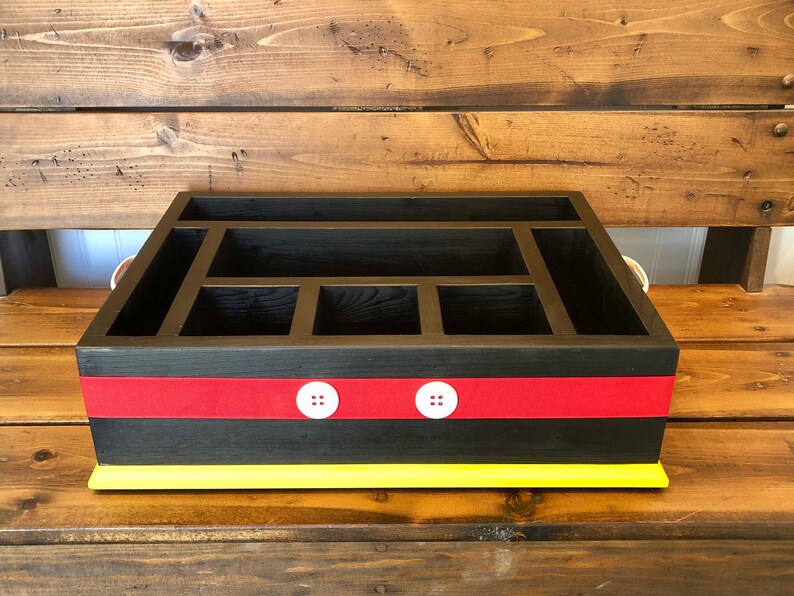 Puede incluir: Un organizador de madera negro con m&uacute;ltiples compartimentos. Presenta una franja roja con detalles de botones blancos y una base amarilla. El organizador est&aacute; dise&ntilde;ado para el almacenamiento y la organizaci&oacute;n.