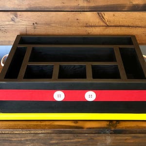 Puede incluir: Organizador de madera negro con m&uacute;ltiples compartimentos, una franja roja con dos botones blancos y una base amarilla. El organizador tiene dos asas color crema y est&aacute; sobre un fondo de madera.