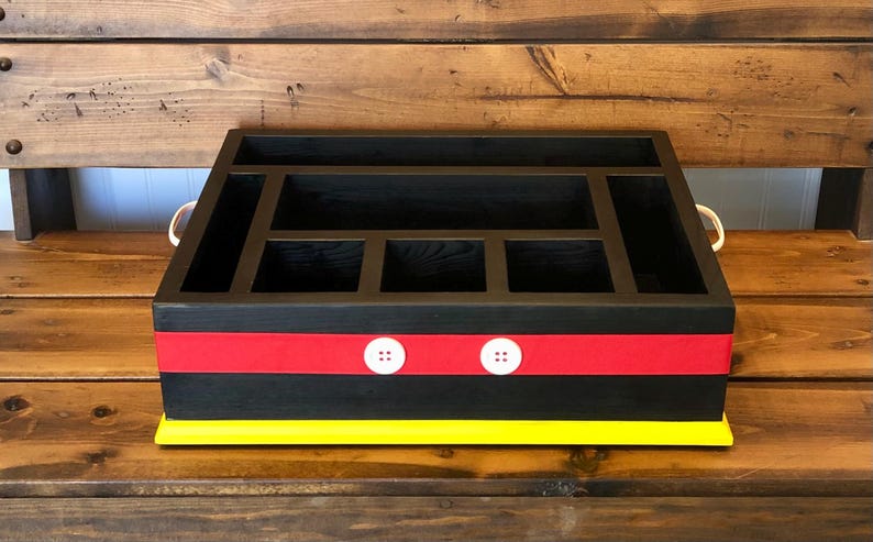 Puede incluir: Organizador de madera negro con m&uacute;ltiples compartimentos, una franja roja con dos botones blancos y una base amarilla. El organizador tiene dos asas color crema y est&aacute; sobre un fondo de madera.