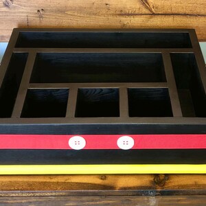 Puede incluir: Organizador de madera negro con m&uacute;ltiples compartimentos. Cuenta con una cinta roja con detalles de botones blancos y una base amarilla. Incluye asas blancas. Ideal para la organizaci&oacute;n de escritorio.