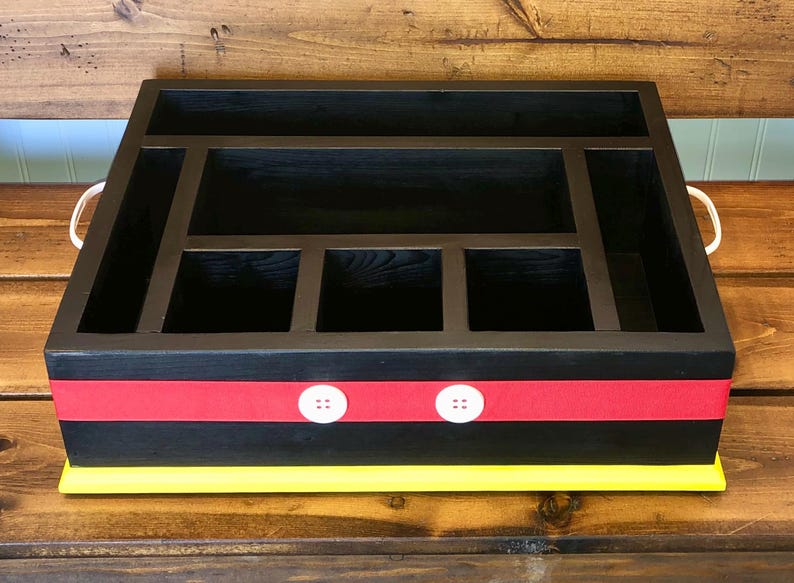 Puede incluir: Organizador de madera negro con m&uacute;ltiples compartimentos. Cuenta con una cinta roja con detalles de botones blancos y una base amarilla. Incluye asas blancas. Ideal para la organizaci&oacute;n de escritorio.