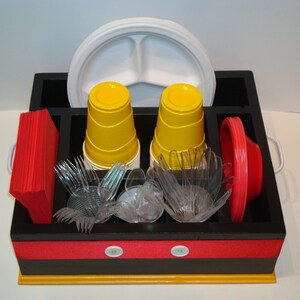 Puede incluir: Un organizador de madera negro y amarillo con compartimentos para vajilla desechable. Contiene vasos amarillos, tenedores y cuencos de pl&aacute;stico transparente, servilletas y platos rojos, y un plato blanco dividido. El organizador tiene una franja roja con botones blancos.