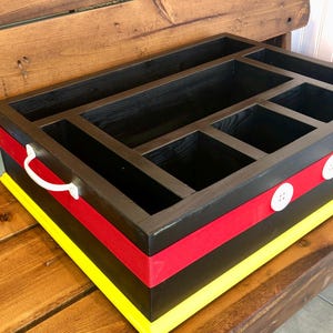 Puede incluir: Un organizador de madera negro con m&uacute;ltiples compartimentos. Cuenta con una cinta roja y un borde amarillo, con botones blancos y un asa. El dise&ntilde;o recuerda a un famoso personaje de dibujos animados.