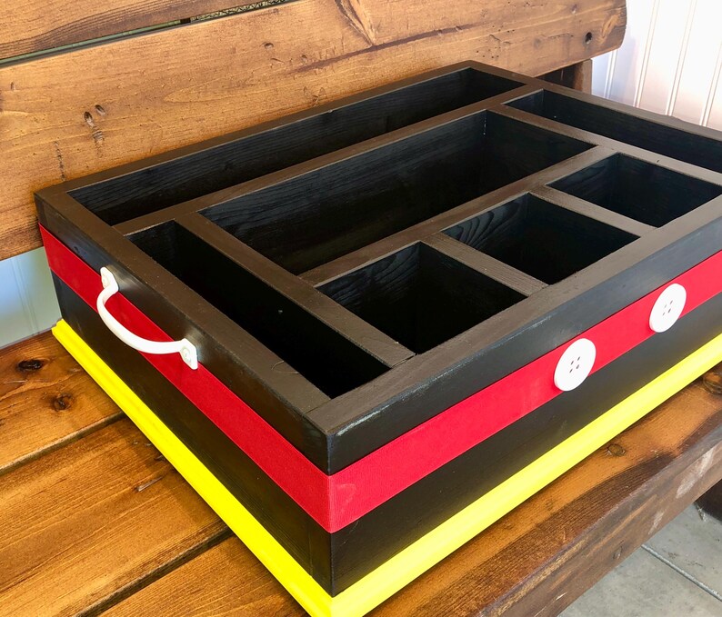 Puede incluir: Un organizador de madera negro con m&uacute;ltiples compartimentos. Cuenta con una cinta roja y un borde amarillo, con botones blancos y un asa. El dise&ntilde;o recuerda a un famoso personaje de dibujos animados.