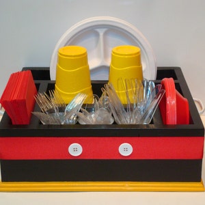 Puede incluir: Un organizador de picnic de madera negra y roja con detalles amarillos, que contiene vasos amarillos, tenedores de pl&aacute;stico transparente, servilletas rojas y un plato blanco dividido. El organizador tiene detalles de botones blancos y asas.