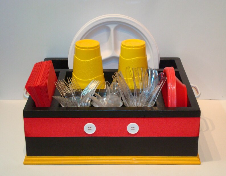 Puede incluir: Un organizador de picnic de madera negra y roja con detalles amarillos, que contiene vasos amarillos, tenedores de pl&aacute;stico transparente, servilletas rojas y un plato blanco dividido. El organizador tiene detalles de botones blancos y asas.