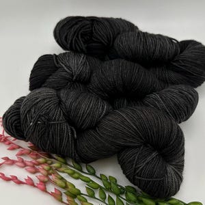 May include: Several skeins of black yarn are arranged on a white surface, with a few sprigs of pink and green decorative foliage. The yarn appears soft and is tightly wound into neat bundles, ideal for knitting or crochet projects.
