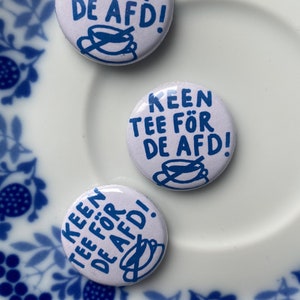 May include: Three white round buttons with blue text and illustrations of teacups. The buttons say "Keen tee for de AFD!"