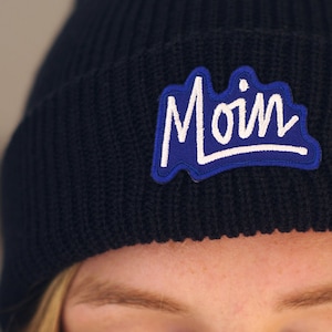 Könnte beinhalten: Eine marineblaue Mütze mit einem blauen und weißen gestickten Aufnäher, auf dem "Moin" steht.