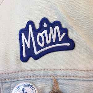 May include: Blue and white embroidered patch with the word "Moin" in white lettering on a blue background. The patch is sewn onto a light blue denim jacket.