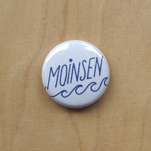 Może przedstawiać: Biały guzik z niebieskim tekstem "Moin sen" i niebieskimi falistymi liniami pod spodem.