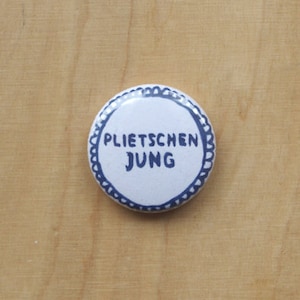 Könnte beinhalten: Ein weißer Knopf mit blauem Rand und dem Text "Plietschen Jung" in Schwarz.