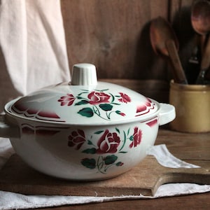 Pode incluir: Prato de servir de cerâmica branca com tampa, com um design floral de rosas vermelhas e folhas verdes. O prato tem duas alças nas laterais.