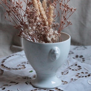 May include: A white ceramic vase with a pedestal base and a small handle. The vase is filled with dried flowers and grasses in shades of beige and brown. The vase sits on a white lace tablecloth.