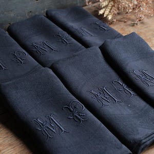 Può includere: Un set di sei tovaglioli di lino blu navy, ciascuno ricamato con un monogramma. I tovaglioli sono piegati con cura e disposti su una superficie di legno. Le iniziali ricamate sono in un carattere decorativo.
