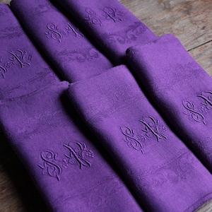 May include: A set of six folded purple linen napkins. Each napkin features an embroidered monogram and decorative floral pattern. The napkins are arranged on a wooden surface, showcasing their rich color and texture.