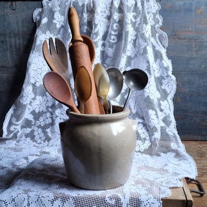 Könnte beinhalten: Eine Sammlung antiker Küchenutensilien, darunter Holzlöffel, ein Nudelholz und Metallkellen, wird in einem beigefarbenen Keramiktopf präsentiert. Die Utensilien sind vor dem Hintergrund einer weißen Spitzendecke und einer rustikalen Holzoberfläche angeordnet.