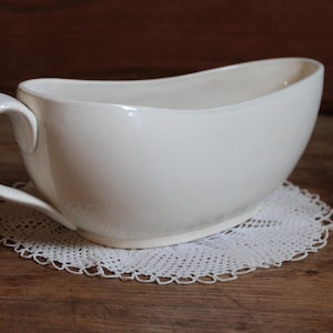 May include: A white ceramic gravy boat with a curved handle and a wide, oval shape. It sits on a white lace doily, with a wooden background. The gravy boat is a classic piece of tableware.