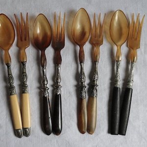 May include: A selection of antique spoons and forks. The utensils feature light brown, horn-like bowls and tines, with ornate silver-coloured metal and ivory or dark brown handles. The spoons and forks are arranged on a white surface.