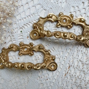 May include: Two ornate gold-toned metal drawer pulls with a decorative scroll design. The pulls have a rectangular shape with a curved handle.