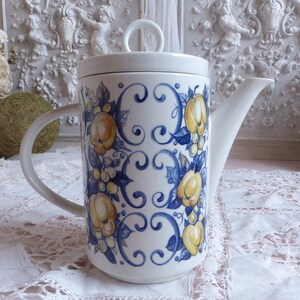 Puede incluir: Tetera de cerámica blanca con un diseño floral azul y amarillo. La tetera tiene una tapa y un asa.