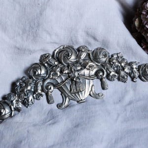 Peut inclure: Une pièce décorative en métal argenté en forme de vase avec un motif floral. La pièce est composée de plusieurs roses et feuilles, toutes finement détaillées.