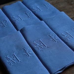 Può includere: Sei tovaglioli di lino blu con iniziali ricamate. Ogni tovagliolo presenta la lettera "M" e un'altra lettera, forse "R", in una scrittura decorativa. I tovaglioli sono piegati con cura e disposti su una superficie di legno.