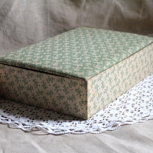 Puede incluir: Una caja rectangular con tapa con bisagras, cubierta con un estampado floral verde claro. La caja está sobre un tapete de encaje blanco. Una flor de peonía rosa y crema está en el fondo.