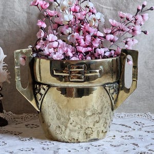 May include: A gold-coloured metal vase with a hammered texture, holding pink and white flowers. The vase has a unique design with handles and decorative details. The arrangement sits on a white lace tablecloth, creating a vintage aesthetic.
