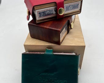 Puede incluir: Tres estuches de cartas de cuero apilados uno encima del otro. El estuche superior es rojo, el estuche del medio es marrón y el estuche inferior es verde. El estuche inferior tiene el texto "The Shadowgan" grabado en la parte delantera.