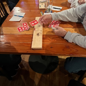 May include: A wooden cribbage board with a red and white playing card on top of it. There are several other playing cards on the table, as well as a glass of beer and a pen.