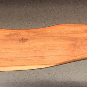 May include: A wooden spatula with a curved handle and a rounded tip. It is made of light brown wood and has the text "MADE FOR GAYLE LYNN WWW.SAWTEQ.COM 05.20.2019" engraved on the back.