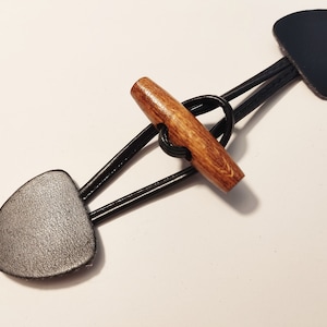Könnte beinhalten: Ein schwarzer Leder-Toggle-Verschluss mit einem Holz-Toggle. Der Toggle ist braun und hat eine glatte Oberfläche.