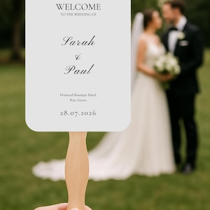 May include: A wedding fan with a light wooden handle and a white card. The card reads "WELCOME TO THE WEDDING OF Sarah & Paul" with the venue and date: Diamond Boutique Hotel, Kos, Greece, 28.07.2026. A couple is blurred in the background.
