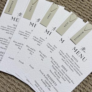 May include: A stack of white wedding menu cards with a minimalist design, held together by silver paper clips. Each menu features a list of dishes and guest names. The menu includes mains, sides, and desserts.