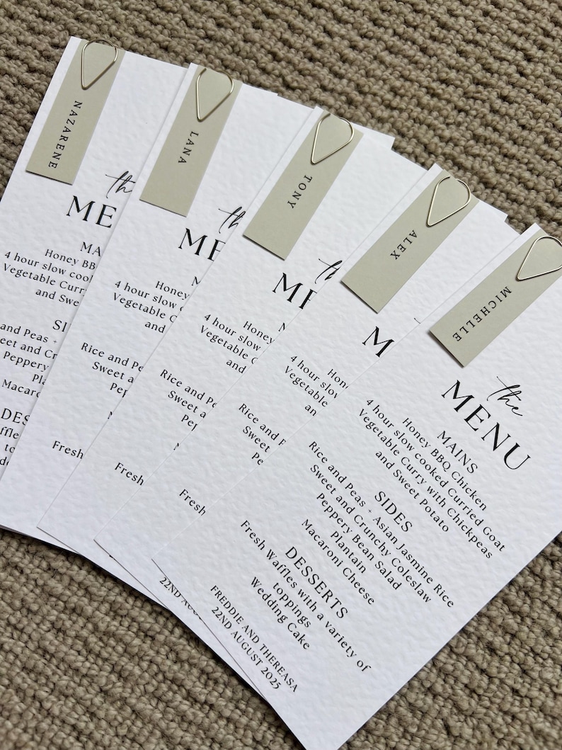 May include: A stack of white wedding menu cards with a minimalist design, held together by silver paper clips. Each menu features a list of dishes and guest names. The menu includes mains, sides, and desserts.