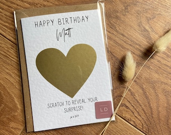 May include: A white birthday greeting card in a clear plastic sleeve featuring the text HAPPY BIRTHDAY MATT in black ink, a large gold heart-shaped scratch-off area, and the text SCRATCH TO REVEAL YOUR SURPRISE! XOXO with a small pink square logo.