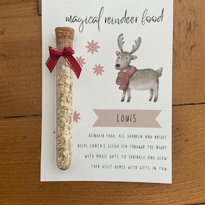 May include: A glass test tube filled with oats, sealed with a cork and a red ribbon, attached to a card featuring a reindeer illustration, snowflakes, the name Louis, and the text: magical reindeer food, reindeer food, all sparkly and bright.