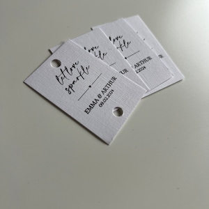 May include: A stack of white gift tags with black calligraphy that reads "let love sparkle" and "love like" with the text "Emma & Arthur 09.02.2024" on the bottom tag.