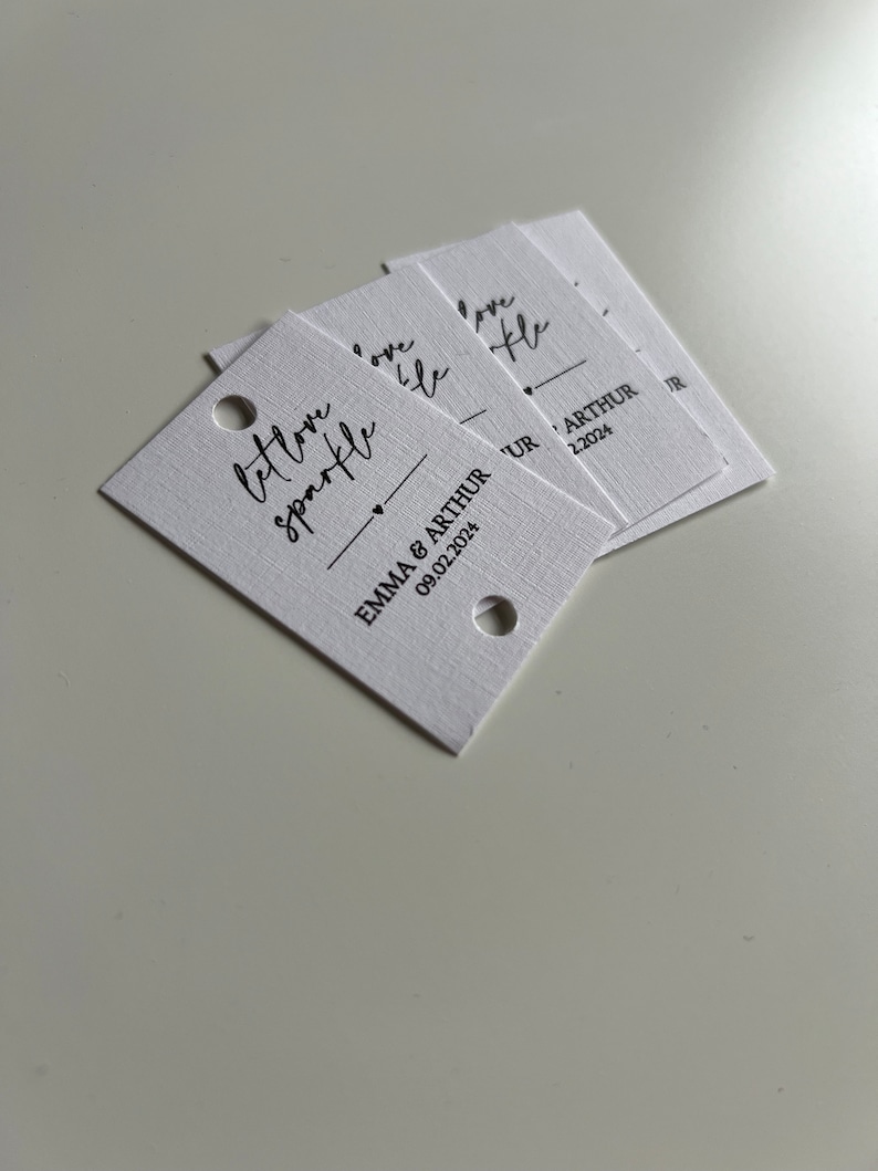 May include: A stack of white gift tags with black calligraphy that reads "let love sparkle" and "love like" with the text "Emma & Arthur 09.02.2024" on the bottom tag.